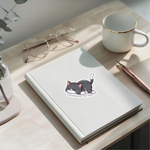 Puede incluir: Una pegatina blanca con un gato de dibujos animados en tonos grises, negros y blancos, tumbado con los ojos cerrados. El texto "OVERTHINKING BUT ALSO HUNGRY" está debajo. La pegatina está sobre un cuaderno blanco.