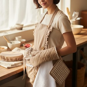 May include: A beige apron with ruffle trim and large wooden buttons. The apron is paired with a matching oven mitt, pot holder, and a white towel. Other kitchen accessories include a bowl cover and a drawstring bag. The items are on a wooden countertop.
