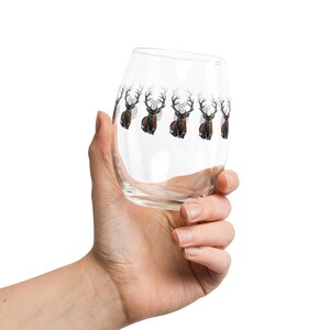 May include: A clear stemless wine glass with a repeating pattern of brown deer with large antlers. The glass is held in a hand against a white background. The deer are depicted in a watercolor style.