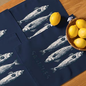 May include: Navy blue placemats with a white sardine illustration and the word "Sardines". The placemats are arranged on a wooden surface with lemons and a woven basket.