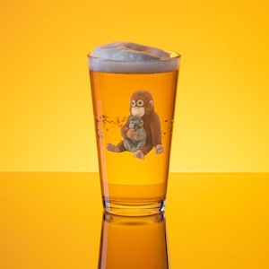May include: A pint glass filled with beer and a foamy head. The glass features a cartoon illustration of a brown orangutan holding a smaller gray monkey. The background is a gradient of yellow.