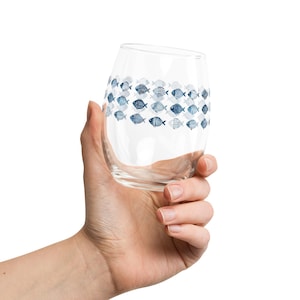 May include: A clear stemless wine glass with a decorative band of blue fish illustrations. The glass is held in a hand against a white background. The fish designs vary in pattern and shade of blue.
