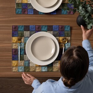 May include: Overhead shot of a wooden table set for two. Each place setting includes a white plate, a smaller plate, a gold fork and spoon, and a colorful patchwork placemat. A person's hands are visible, and a black vase with greenery is on the table.
