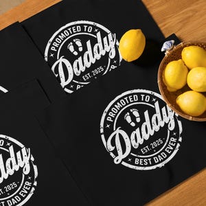 May include: Black aprons with a white circular design that reads "PROMOTED TO Daddy EST. 2025 BEST DAD EVER" and baby footprints. A basket of lemons and two loose lemons are also visible.