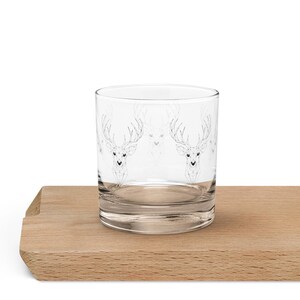 May include: A clear glass tumbler with a repeating pattern of stylized deer heads. The deer are depicted in a geometric, line-art style. The glass sits on a light-colored wooden surface.