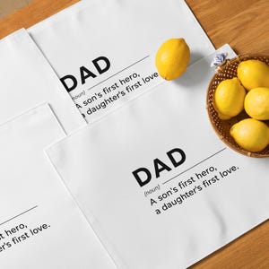 May include: White cotton kitchen towels with the word "DAD" printed in black, along with the definition "A son's first hero, a daughter's first love." A basket of yellow lemons sits nearby.
