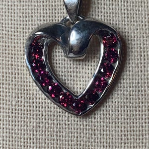 May include: A silver heart-shaped pendant necklace with a small, twisted silver loop at the top and a border of small, dark red gemstones.