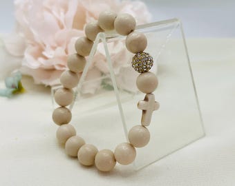 Op de afbeelding: Een beige kralen armband met een witte kruis bedel en een gouden strass bal bedel.