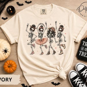 Puede incluir: Una camiseta de manga corta color marfil con un gráfico de cuatro esqueletos bailando. Los esqueletos llevan accesorios como diademas, un tutú y un sombrero, y están representados con varias zapatillas y botas. Pequeños corazones rosas y estrellas rodean a las figuras.