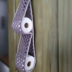 May include: A crocheted toilet paper holder in a purple and white color scheme hangs on a wooden door. Two rolls of toilet paper are held in the holder.