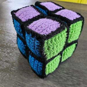 May include: A crocheted cube toy with black, green, blue, and purple squares. The cube is made of yarn and has a soft, textured surface.