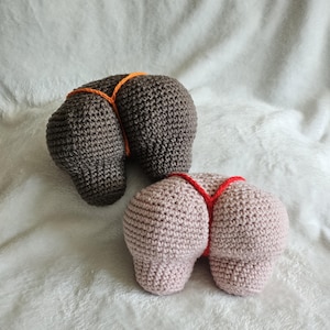 May include: Two crocheted butt-shaped items. One is brown with an orange string, and the other is pink with a red string. The items are on a white, textured surface.
