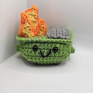 May include: A green crocheted trash can with a smiling face and black eyes. The trash can has a gray lid and orange flames on top.