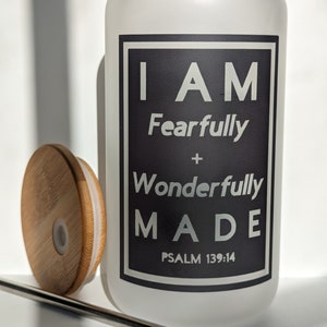 May include: A white frosted glass water bottle with a black square label that reads "I AM Fearfully + Wonderfully MADE PSALM 139:14". The bottle has a bamboo lid and a metal straw.
