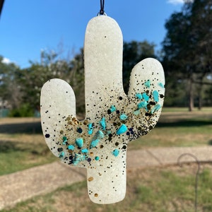 May include: White cactus-shaped ornament with gold and turquoise glitter and embellishments. The ornament is hanging from a black string.