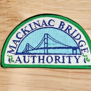 May include: A semi-circular embroidered patch featuring a light blue center with a dark blue outline of the Mackinac Bridge. The white border contains the dark blue text MACKINAC BRIDGE AUTHORITY and small green accents, finished with a green edge.