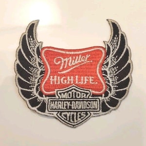 Puede incluir: Un parche bordado que presenta un emblema rectangular rojo con el texto blanco Miller High Life, colocado sobre un logotipo de escudo en blanco y negro de Harley-Davidson Motor Cycles, flanqueado por dos grandes alas emplumadas en blanco y negro.