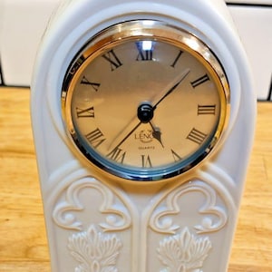 May include: A cream-colored ceramic clock with a gold-tone clock face and Roman numerals. The clock has a decorative, arched shape with embossed floral designs. The clock face reads "LENOX QUARTZ".