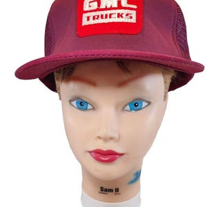 May include: A burgundy trucker hat with a mesh back and a curved brim. The front features a red square patch with the white text "GMC TRUCKS". The hat is displayed on a mannequin head with blue eyes and red lips.