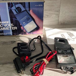 May include: A Micronta 7-Range AC Volt-Ammeter clamp meter with a black plastic body, an analog display, and a red and black test lead set. The device is accompanied by its original cardboard packaging box and a printed instruction manual.
