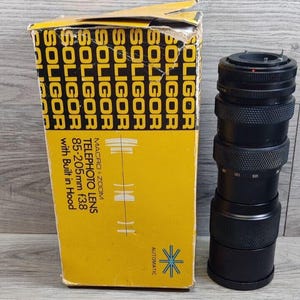May include: A black Soligor Macro Zoom Telephoto Lens 85-205mm f3.8 with a yellow and black box. The box has the Soligor logo repeated and the words "Macro+Zoom Telephoto Lens 85-205mm f3.8 with Built in Hood". The lens has measurement markings.