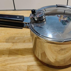 以下が含まれることがあります： 黒いハンドルと光沢のある反射性の蓋が付いたステンレス製の圧力鍋。鍋は木製の表面に置かれ、蓋には圧力解放バルブが付いています。