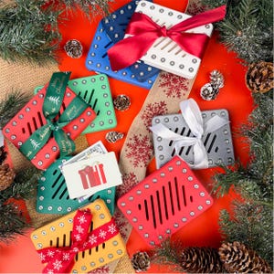 May include: A festive arrangement of colorful, slotted gift boxes in red, green, yellow, blue, and white, each adorned with ribbons. The boxes are surrounded by pine cones, greenery, and a decorative ribbon, suggesting a holiday theme.
