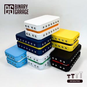May include: A stack of six small, rectangular boxes with different colored lids. The boxes are made of a textured material and have a row of silver screws around the edges. The boxes are labeled "Binary Garage" and "Infuariating".