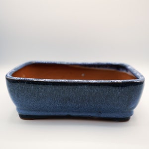 May include: A rectangular bonsai pot with a dark blue exterior and a brown interior. The pot has a slightly textured surface and a dark rim. The base of the pot is a darker shade of blue.