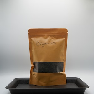 May include: A brown paper bag with the word "Organics" written on it. The bag is filled with black seeds and is sitting on a black plastic tray.