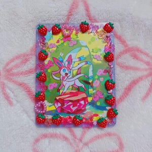 May include: A clear rectangular case with a colourful illustration of a Sylveon character. The case is decorated with red strawberry charms and decorative accents. The background is a soft pink.