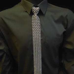 May include: A silver chainmail tie is shown against a dark green shirt. The tie is constructed from small, interlocking metal rings, giving it a textured, metallic look. The tie's design is a unique alternative to a traditional tie.