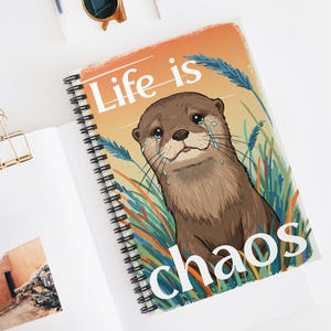 May include: A spiral-bound notebook with a cover featuring a cartoon otter with tears, surrounded by grass, and the words "Life is chaos". The notebook is open on a white surface with a gold binder clip.