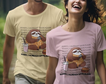 Pode incluir: Duas pessoas vestindo camisetas com um gráfico de um bicho-preguiça e o texto "Desculpe, já tenho planos". O bicho-preguiça está segurando um telefone e um milkshake. As pessoas estão sorrindo e caminhando em uma floresta.