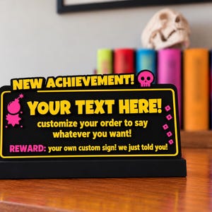 May include: A black rectangular desk sign with yellow and pink text that reads NEW ACHIEVEMENT! with a skull icon, YOUR TEXT HERE!, customize your order to say whatever you want!, and REWARD: your own custom sign! we just told you!.