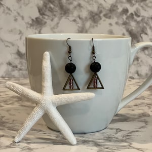 Könnte beinhalten: Ein Paar Hängeohrringe mit schwarzen Lavasteinen und einem Dreiecksdesign. Das Dreieck ist aus Metall mit einer Bronze-Oberfläche. Die Perlen sind auf einem Draht aufgefädelt und hängen am Dreieck. Die Ohrringe liegen auf einer weißen Oberfläche.
