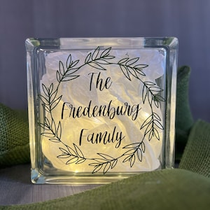May include: A clear glass block with a white background and a black wreath design. The text "The Fredenburg Family" is inside the wreath.