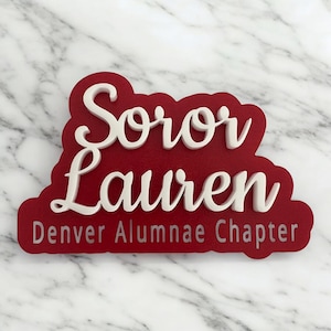 May include: A red sign with white lettering that reads "Soror Lauren Denver Alumnae Chapter." The sign has a layered design, with the white letters raised above the red base. The sign is likely for a sorority.