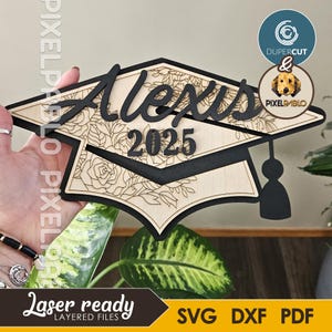 Könnte beinhalten: Eine Holz-Abschluss-Kappe mit dem Namen "Alexis" und dem Jahr "2025" eingraviert. Die Kappe ist mit einem floralen Design verziert und hat einen schwarzen Quasten.