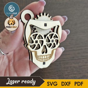 Puede incluir: Un colgante de calavera de madera cortada con láser con un diseño de engranaje. La calavera presenta intrincados recortes para los ojos y los dientes, con un engranaje en la parte superior. El texto "Laser ready" y las opciones de tipo de archivo son visibles.