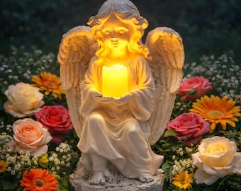 May include: A cream-colored angel figurine with detailed wings and flowing robes, kneeling while holding a glowing, warm yellow LED candle. The sculpture features a serene facial expression and is surrounded by various colorful flowers.