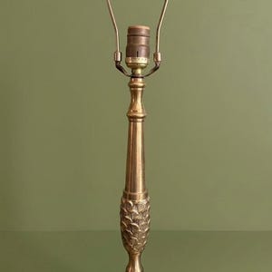 May include: A brass table lamp base featuring a circular, textured base with a radiating leaf pattern. The central column includes a decorative section with carved leaf details, a smooth tapered stem, and a metal harp attachment at the top.