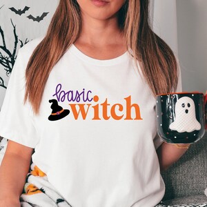 Pode incluir: Camiseta branca com o texto "basic witch" em laranja e roxo. Um chapéu de bruxa é representado em roxo com uma borda preta. A camiseta é usada por uma pessoa sentada em um sofá com uma manta de padrão preto e branco.