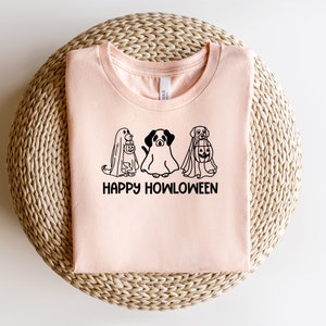 K&ouml;nnte beinhalten: Ein pinkfarbenes T-Shirt mit einer schwarzen Grafik von drei Hunden, die als Geister f&uuml;r Halloween verkleidet sind. Der Text "Happy Howloween" ist unter den Hunden gedruckt.