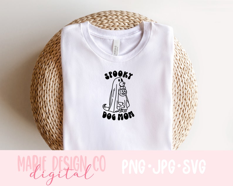 Op de afbeelding: Wit T-shirt met een zwart-witte afbeelding van een hond die verkleed is als een geest en een pompoen vasthoudt. De tekst "Spooky Dog Mom" staat onder de afbeelding.