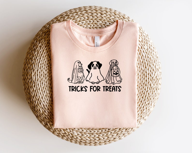 K&ouml;nnte beinhalten: Ein pinkfarbenes T-Shirt mit einer schwarzen Grafik von drei Hunden, die als Geister f&uuml;r Halloween verkleidet sind. Der Text "TRICKS FOR TREATS" ist unter der Grafik gedruckt.