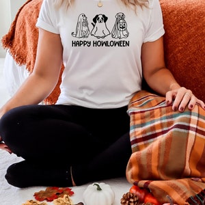 K&ouml;nnte beinhalten: Ein wei&szlig;es T-Shirt mit einer schwarzen Grafik von drei Hunden, die als Geister f&uuml;r Halloween verkleidet sind. Der Text "HAPPY HOWLOWEEN" ist unter der Grafik gedruckt.