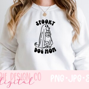 Op de afbeelding: Wit sweatshirt met een zwart-witte illustratie van een hond die verkleed is als een geest en een pompoen vasthoudt. De tekst "Spooky Dog Mom" staat onder de illustratie.