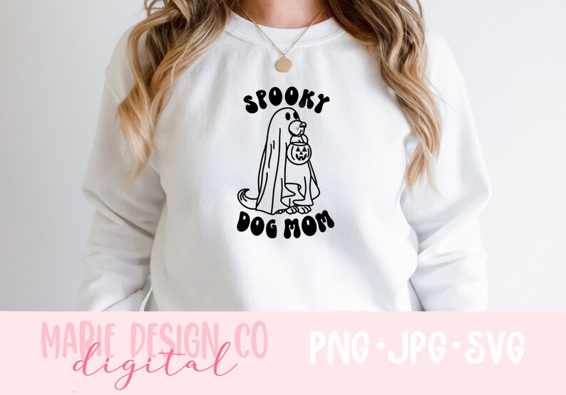 Op de afbeelding: Wit sweatshirt met een zwart-witte illustratie van een hond die verkleed is als een geest en een pompoen vasthoudt. De tekst "Spooky Dog Mom" staat onder de illustratie.