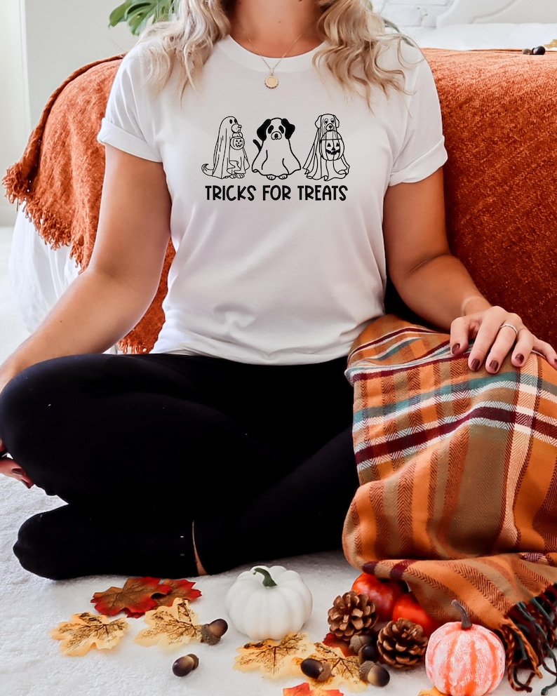 K&ouml;nnte beinhalten: Ein wei&szlig;es T-Shirt mit einer schwarzen Grafik von drei Hunden, die als Geister verkleidet sind. Die Hunde halten K&uuml;rbisse und der Text "TRICKS FOR TREATS" befindet sich unter der Grafik.